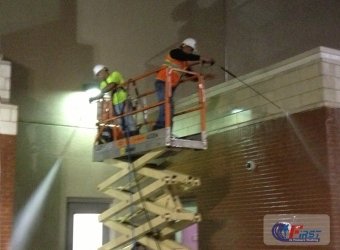 Two workers in hard hats and safety vests perform commercial pressure washing on a brick building using a scissor lift.